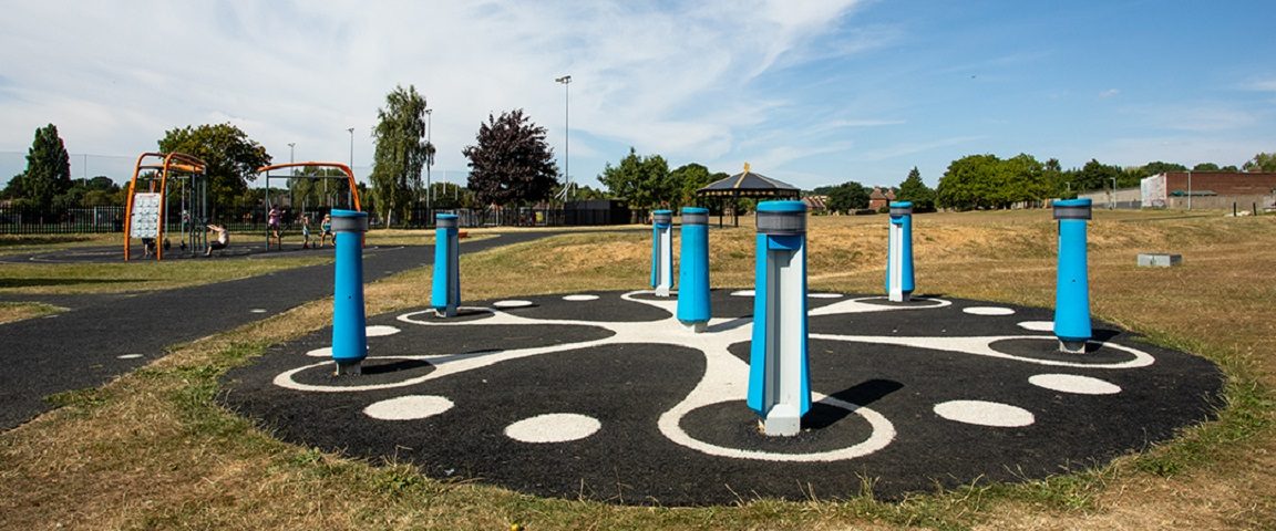 grassy area with black tarmac decorated with a white swirl and dot pattern. There are seven blue poles with a black band around the top.